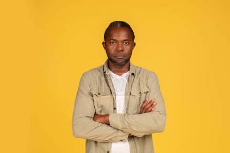 Téléchargez les photos : Serious confident middle aged african american male in casual crossed arms on chest, looking at camera, isolated on yellow background, studio. Lifestyle, emotions, discontent, conflict and negative - en image libre de droit