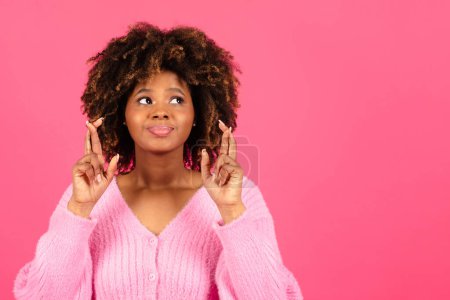 Foto de Cheerful millennial african american curly lady in casual crossed fingers, make wish isolated on pink background, studio. Belive gesture, hope for success, desire, ad and offer, fashion and lifestyle - Imagen libre de derechos