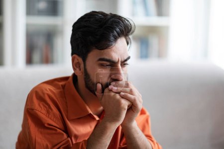 Photo for Closeup photo of sad depressed attractive bearded young middle eastern man in orange shirt sitting on couch at home, covering his mouth with hands, thinking about something, copy space - Royalty Free Image