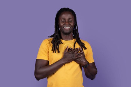 Téléchargez les photos : Peaceful smiling young african american guy in yellow t-shirt with closed eyes holding hands on chest, expressing love, gratitude, appreciation, purple studio background. Human gestures concept - en image libre de droit