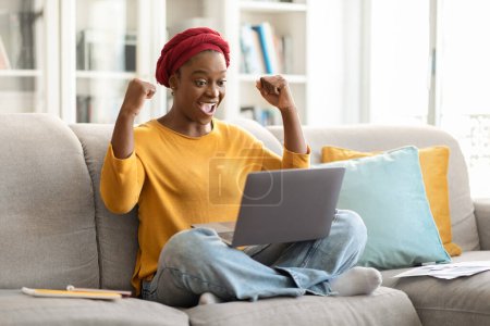 Téléchargez les photos : Emotional cheerful young black lady in casual wearing red headscarf entrepreneur sitting on couch with legs up, using laptop and gesturing, celebrating success, made good business deal, copy space - en image libre de droit