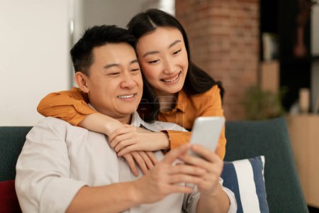Photo for Happy asian man and woman sitting on couch, using smartphone, looking at cellphone screen and smiling, using newest mobile application, home interior, copy space - Royalty Free Image