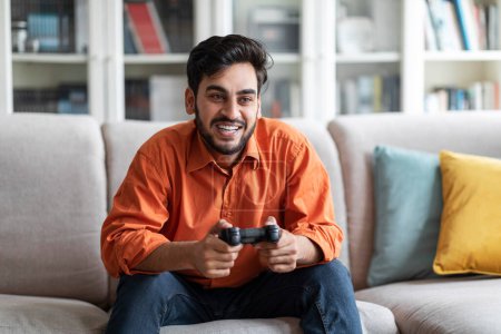 Photo for Resting at home, domestic entertainment. Handsome young middle eastern man playing video games and laughing while sitting alone on the sofa at home, free space. Modern technologies and entertainment - Royalty Free Image