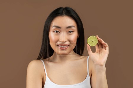 Foto de Happy young japanese lady with clean skin, holding lime half near her face and smiling at camera, posing with citrus fruit over brown background, studio shot - Imagen libre de derechos
