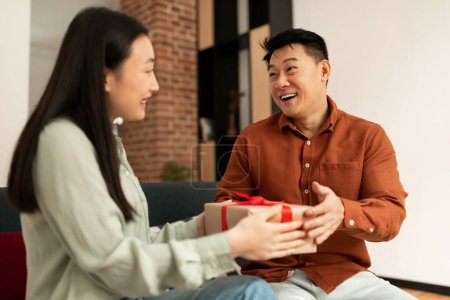 Foto de Loving young asian wife giving present to her happy mature husband, sitting on couch at home. Positive woman greeting spouse with birthday or anniversary, surprising with gift box - Imagen libre de derechos