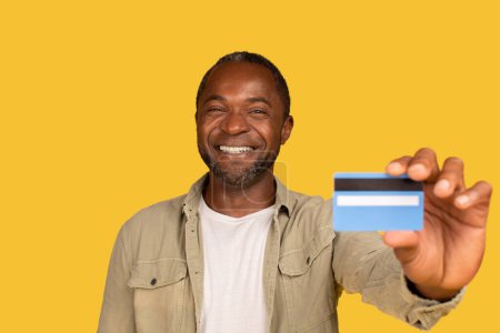 Téléchargez les photos : Glad middle aged african american male in casual show credit card to check bank financial account, recommend money isolated on yellow background, studio. Buy, online shopping, cashback, ad and offer - en image libre de droit
