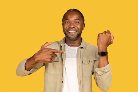 Téléchargez les photos : Happy middle aged african american man in casual show finger at smartwatch with blank screen, isolated on yellow background, studio. Health monitoring, gadget approving, recommendation, ad and offer - en image libre de droit