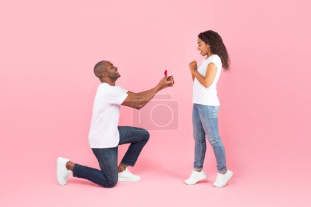 Téléchargez les photos : Loving black middle aged man standing on one knee and offering engagement ring to his beloved young woman, pink studio background, side view. Male making proposal to sweetheart - en image libre de droit