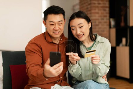 Téléchargez les photos : Happy asian man and woman holding smartphone and credit card, shopping online from home, sitting on sofa. Korean spouses using mobile phone and making purchases - en image libre de droit