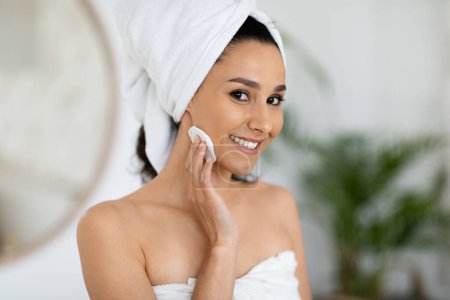 Téléchargez les photos : Cheerful middle eastern millennial curly female in towel after shower cleaning skin with cotton pad in bedroom interior, close up. Good morning at home, daily beauty care and anti-aging treatments - en image libre de droit