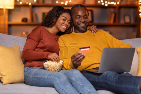 Foto de Relaxed african american spouses happy middle aged man and young woman sitting on couch, eating popcorn, hugging, using computer and credit card at home, planning vacation, booking flights online - Imagen libre de derechos