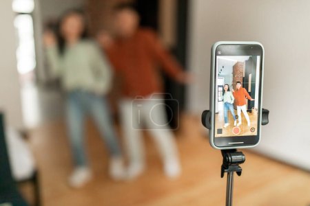 Téléchargez les photos : Asian husband and wife recording video content for social media, dancing in front of cellphone camera, selective focus on smartphone set on tripod, home interior - en image libre de droit