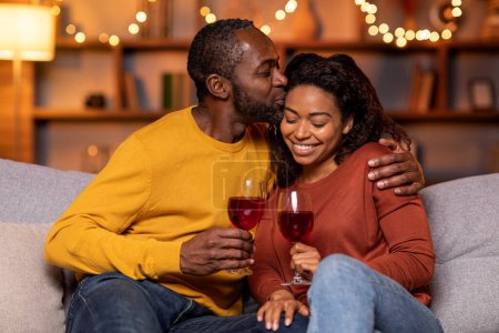 Foto de Cheers. Closeup of beautiful smiling young american couple drinking red wine, holding glasses and toasting, having romantic dinner, spending time together. Young black family celebrating - Imagen libre de derechos