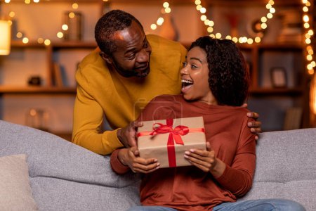 Photo for Happy anniversary my love. Handsome african american man greeting his beloved wife or girlfriend, guy hugging his pretty cheerful woman and giving gift box, festive home interior, copy space - Royalty Free Image