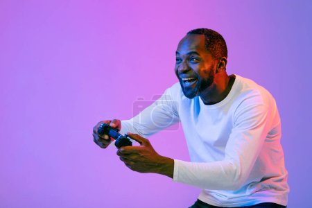Téléchargez les photos : Joyful emotional african american middle aged man playing video games with joystick over neon studio background, looking at copy space and smiling. Video games addiction concept - en image libre de droit