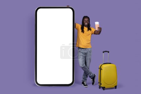 Foto de Happy long-haired young african american guy traveller standing next to smatphone with white empty screen, yellow luggage, showing passport with tickets, mockup, purple studio background - Imagen libre de derechos