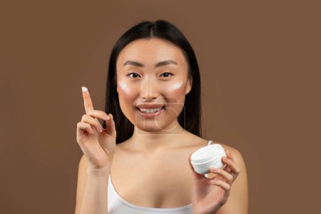 Téléchargez les photos : Happy young japanese lady holding jar of cream and smiling at camera, posing isolated on brown background, studio shot. Cosmetics for beauty care, skin moisturizing, spa, daily beauty treatments - en image libre de droit