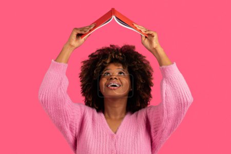 Photo for Happy funny young african american curly woman student with braces lifts book up and reads, search info, isolated on pink background, studio. Literature, study, knowledge in free time and fun alone - Royalty Free Image