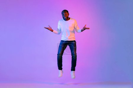 Photo for Positive happy middle aged african american guy in casual outfit jumping in the air and gesturing, raising hands up over neon light studio background, full length shot, copy space - Royalty Free Image