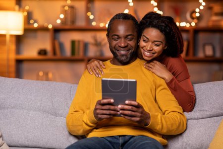 Foto de Happy smiling african american spouses husband and wife sitting on couch, using digital pad at cozy home interior decorated with lights, shopping online, surfing on Internet, copy space - Imagen libre de derechos