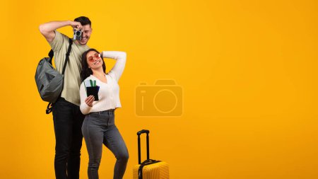 Téléchargez les photos : Cheerful millennial arabic guy takes photo camera and woman in glasses with suitcase and tickets rejoice to trip, isolated on orange background, studio. Travel at vacation, journey and fun together - en image libre de droit