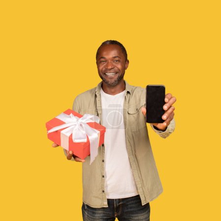Téléchargez les photos : Glad african american middle aged guy show box with gift and smartphone with empty screen isolated on orange background, studio. App for congratulations on holiday and birthday remote, buy present - en image libre de droit