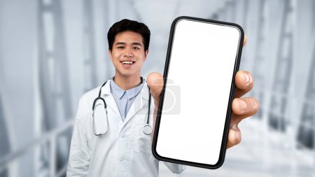 Foto de Smiling Asian Male Doctor Wearing Uniform Showing Big Smartphone With Blank Screen At Camera While Standing In Clinic Hall, Smiling Therapist Man Demonstrating Empty Cellphone, Collage, Mockup - Imagen libre de derechos