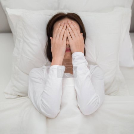 Téléchargez les photos : Top view of unhappy brunette young woman in white pajamas lying in white bed at home, covering her face from morning light with palms. Depression, anxiety, mental health concept - en image libre de droit