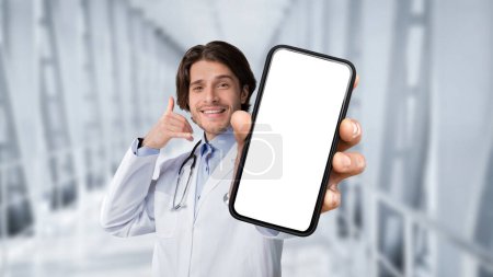Foto de Telemedicine Concept. Smiling Doctor Man Holding Blank Smartphone And Showing Call Me Gesture While Standing In Clinic Hall, Handsome Male Physician Advertising Modern App For Online Medicine, Mockup - Imagen libre de derechos