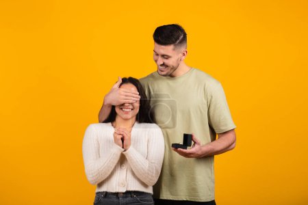 Foto de Happy millennial tall arab guy close eyes to amazed woman, hold box with ring, isolated on orange background, studio, free space. Marriage proposal, surprise betrothal, love, romance and relationship - Imagen libre de derechos