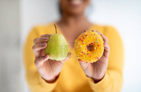 Foto de Smiling young african american lady in yellow clothes show pear and donut in minimalist kitchen interior, close up, blurred. Choice of sweets or healthy food, diet, weight loss, body care at home - Imagen libre de derechos