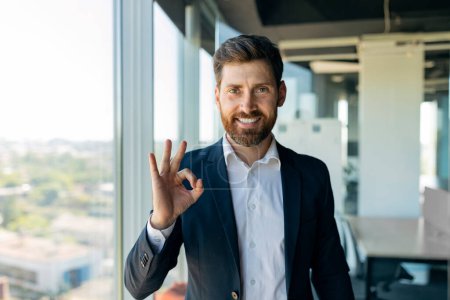 Photo for Cheerful middle aged businessman in formal wear showing ok sign and smiling at camera, standing in office interior. Great deal, business, work, study, recommendation from professional - Royalty Free Image