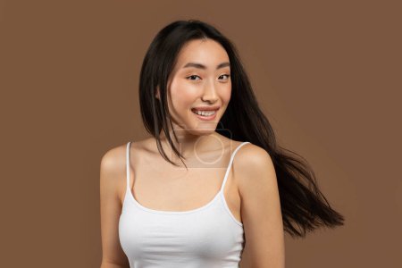 Téléchargez les photos : Portrait of happy korean woman posing with long and healthy flying hair on brown studio background and smiling at camera. Haircare and fashion concept - en image libre de droit