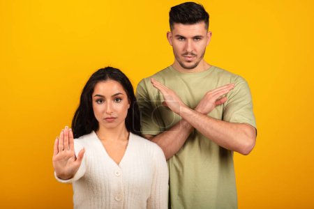 Foto de Serious strict millennial arab guy and woman in casual make stop sign and cross arms, isolated on orange background, studio. Gesture against harassment and discrimination, say no, fight with covid-19 - Imagen libre de derechos