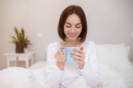 Téléchargez les photos : Happy beautiful brunette young woman in white pajamas sitting in bed, holding blue mug and smiling, cheerful lady drinking coffee in home bedroom after waking up in the morning, copy space - en image libre de droit