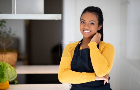Photo for Smiling young african american lady cook in apron near table with vegetables in minimalist kitchen interior. Household chores, prepare healthy food, dinner, professional at work, body care at home - Royalty Free Image