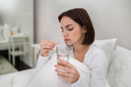 Téléchargez les photos : Close up image of brunette young woman holding glass of water and pink capsule, taking pills to recover from flu or cold, sitting in bed at home. Seasonal disease and medications concept - en image libre de droit
