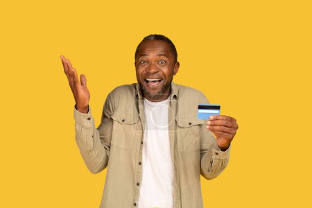 Photo for Cheerful surprised confused middle aged african american man show credit card, raises hand up rejoices to success isolated on yellow background, studio. Win, online shopping and cashback, ad and offer - Royalty Free Image