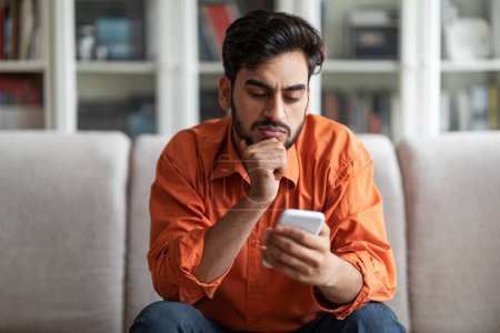 Photo for Thoughtful handsome young middle eastern man sitting on couch alone and using newest phone at home, concentrated arabic guy reading blog or news on Internet, touching his face, copy space - Royalty Free Image