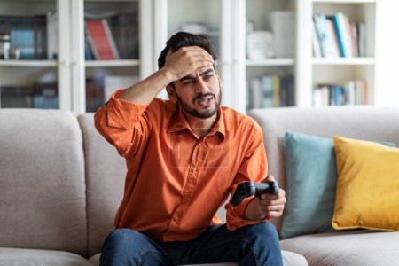 Photo for Sad handsome young arab guy in smart casual playing video games alone at home at weekend, man holding joystick, sitting on couch, touching his forehead, lost in game, copy space - Royalty Free Image