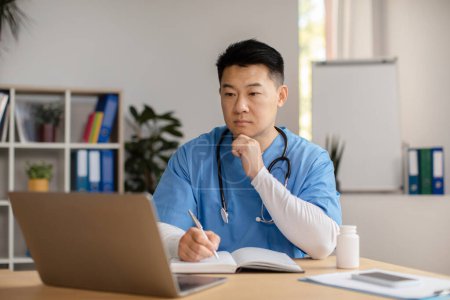 Photo for Pensive serious mature japanese doctor in uniform with stethoscope looks at laptop makes notes at table in clinic office interior. Meeting, support, remote examination, new normal and medical service - Royalty Free Image