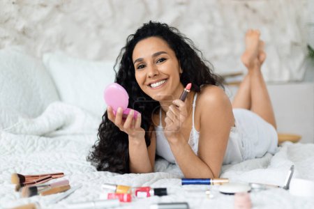 Foto de Smiling pretty arabic millennial curly female in pajamas lies on bed with cosmetics, make makeup, enjoy spare time in bedroom interior. Morning at home, beauty care and daily procedures, nude make-up - Imagen libre de derechos