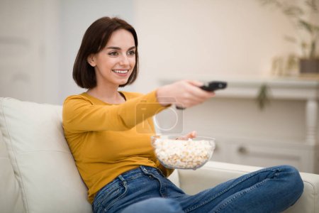 Photo for Cheerful happy attractive young woman sitting on couch in cozy living room, watching TV show, movies with popcorn, holding remote controller, spending time alone at home, copy space - Royalty Free Image