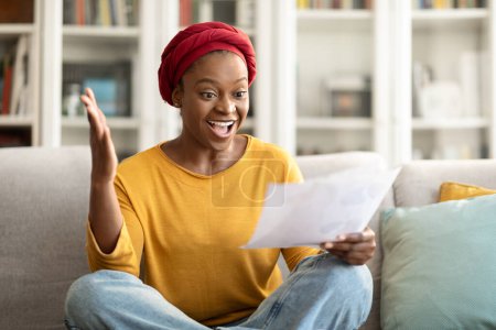 Foto de Happy young african american woman sit on couch at home, reading good news in paper letter checking domestic bills, emotional black lady holding documents doing paperwork, reading correspondence - Imagen libre de derechos