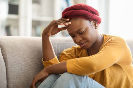 Photo for Lonely depressed unhappy young african american woman wearing red turban sitting on couch with legs up at home, suffering from loneliness, touching her head and crying, copy space - Royalty Free Image