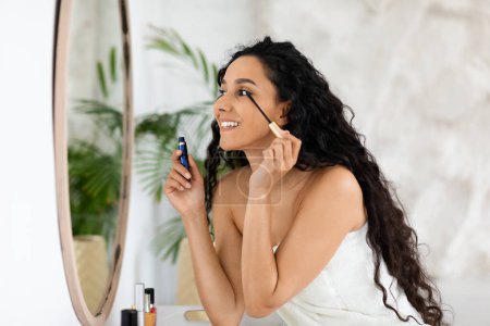 Cheerful arabic millennial curly lady in towel apply mascara on eyelashes, looking in mirror, enjoy nude makeup in bedroom interior, free space. Beauty care, daily treatments, lifestyle and cosmetics