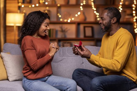 Téléchargez les photos : Loving handsome black man surprising his amazed pretty girlfriend with engagement ring, sitting on the couch at home in the living room decorated with lights. Love, relationships, proposal - en image libre de droit