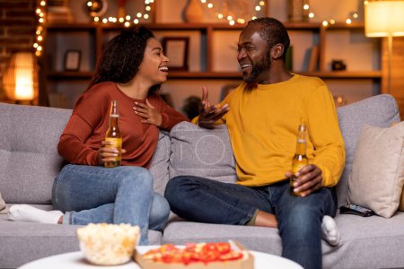 Foto de Black couple in love having date at home, happy cheerful african american young woman and middle aged man sitting on couch, drinking beer, eating pizza, having conversation, laughing, copy space - Imagen libre de derechos