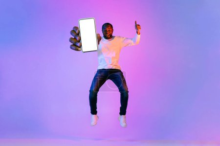 Photo for Excited happy handsome middle aged african american man in stylish casual outfit jumping in the air with brand new smartphone in his hand, showing mockup screen, neon studio background, full length - Royalty Free Image