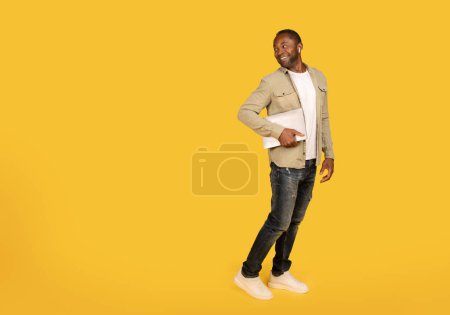 Foto de Glad middle aged african american male in casual, wireless headphones with laptop look back at copy space isolated on yellow background, studio. Gadget for business, freelance and work, ad and offer - Imagen libre de derechos
